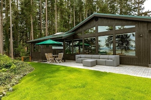 A modern house with a covered patio, a dining area, and a large window overlooking a forest. - Secluded Water & Mountain View Retreat with Sauna (Oak Harbor)