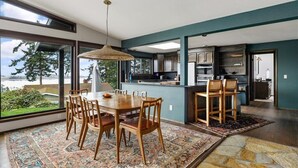 Una cocina moderna con armarios oscuros, un comedor con una gran mesa de madera y sillas, y vistas al exterior a través de amplios ventanales.