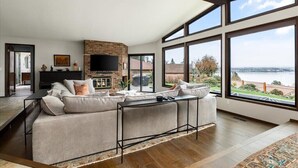 Un salón amplio con un sofá grande, un televisor de pantalla plana y una chimenea. Hay ventanas grandes que ofrecen vistas al exterior.