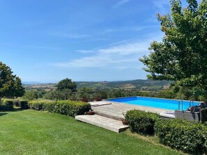 Une piscine dotée d’une terrasse en bois, entourée de pelouse et de haies bien entretenues, et offrant une vue pittoresque sur les collines en arrière-plan.