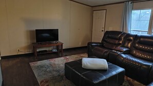 Ein Wohnzimmer mit einem Ledersofa, einem Fernseher auf einem Holzboden und einem kleinen Hocker mit einem Handtuch.