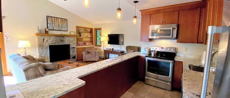 A modern kitchen with a central island, built-in oven, microwave, and refrigerator. There is a seating area with a sofa and a fireplace.