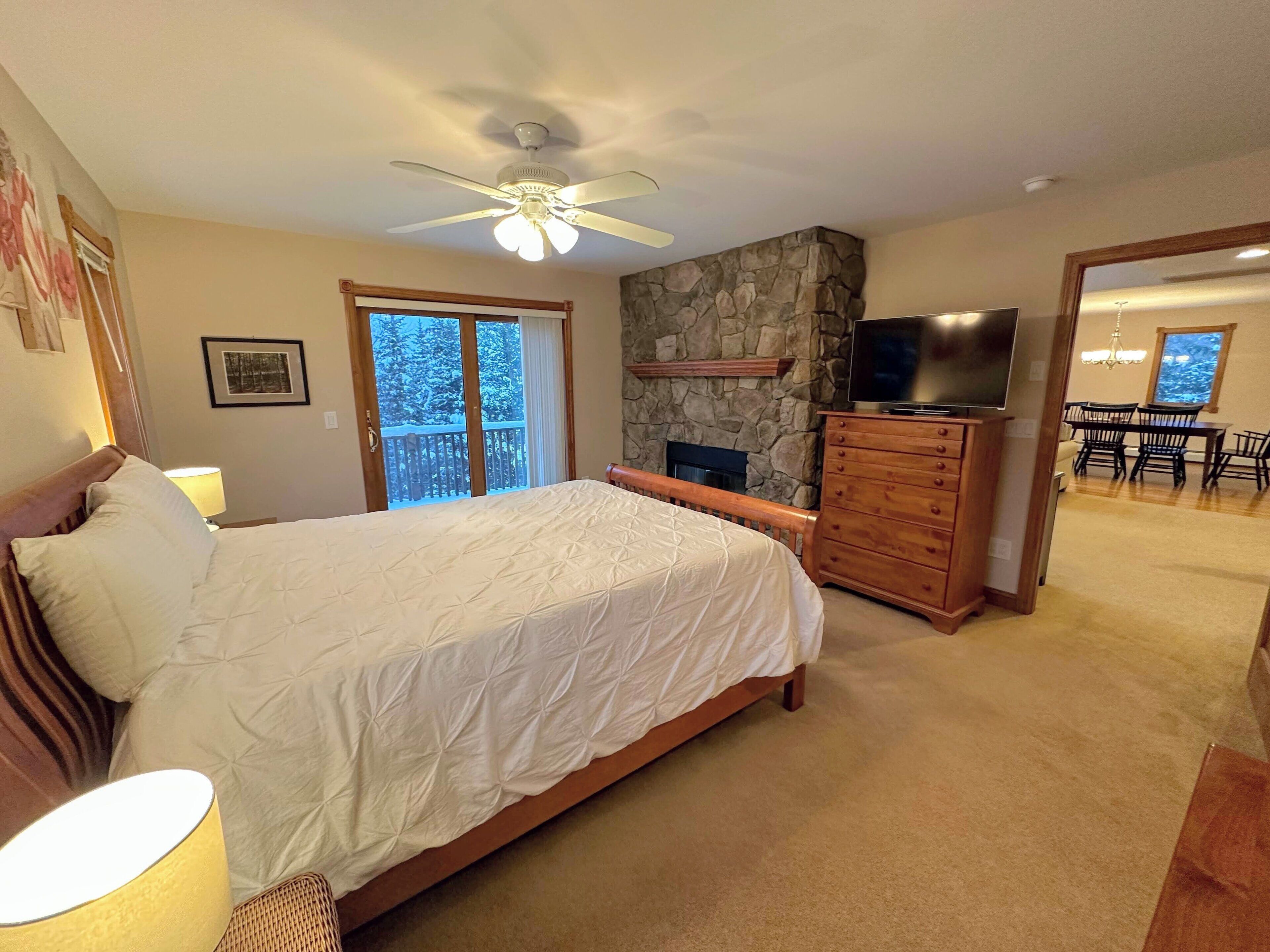 Un dormitorio con cama, cómoda, televisor, chimenea y balcón con vista a árboles.