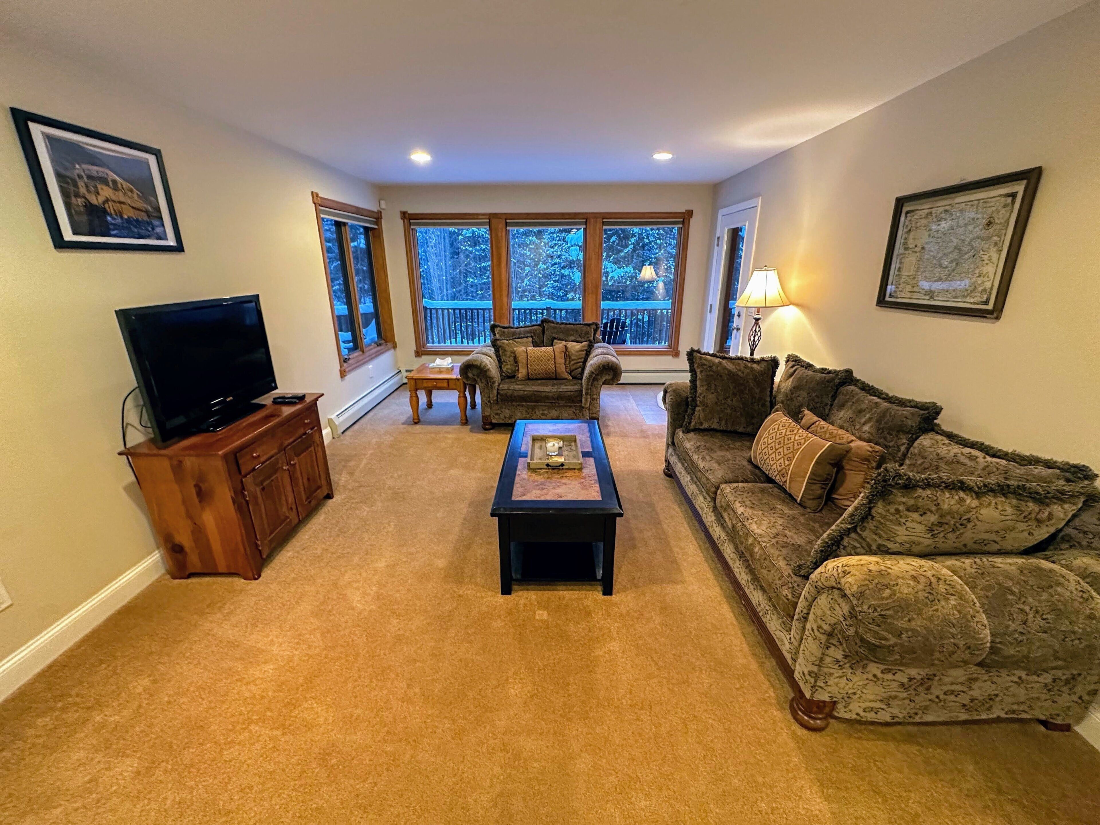 A living room with a television, a coffee table, two sofas, and large windows.