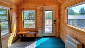 Una cabaña pequeña con paredes de madera y techo inclinado. Hay un banco debajo de una ventana, una puerta con vista a una terraza y un radiador a la derecha.