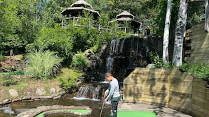 Ein Golf-Putting-Green mit einem kleinen Wasserfall und strohgedeckten Gebäuden im Hintergrund.