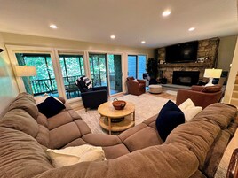 Una sala de estar con un sofá modular, una chimenea, un televisor y un balcón con vistas a árboles.