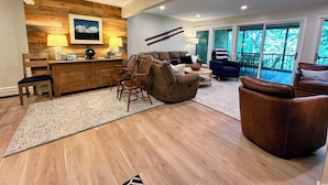 Ein modernes Wohnzimmer mit einer Wand im Holzdesign, einem Sofa, Sesseln, einem Couchtisch und Blick ins Freie durch große Schiebetüren.