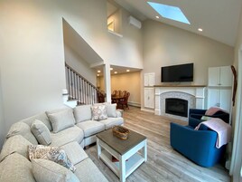 Amplia sala de estar con sofá modular, un sillón azul, chimenea, televisor montado arriba y una escalera que sube a un nivel superior.