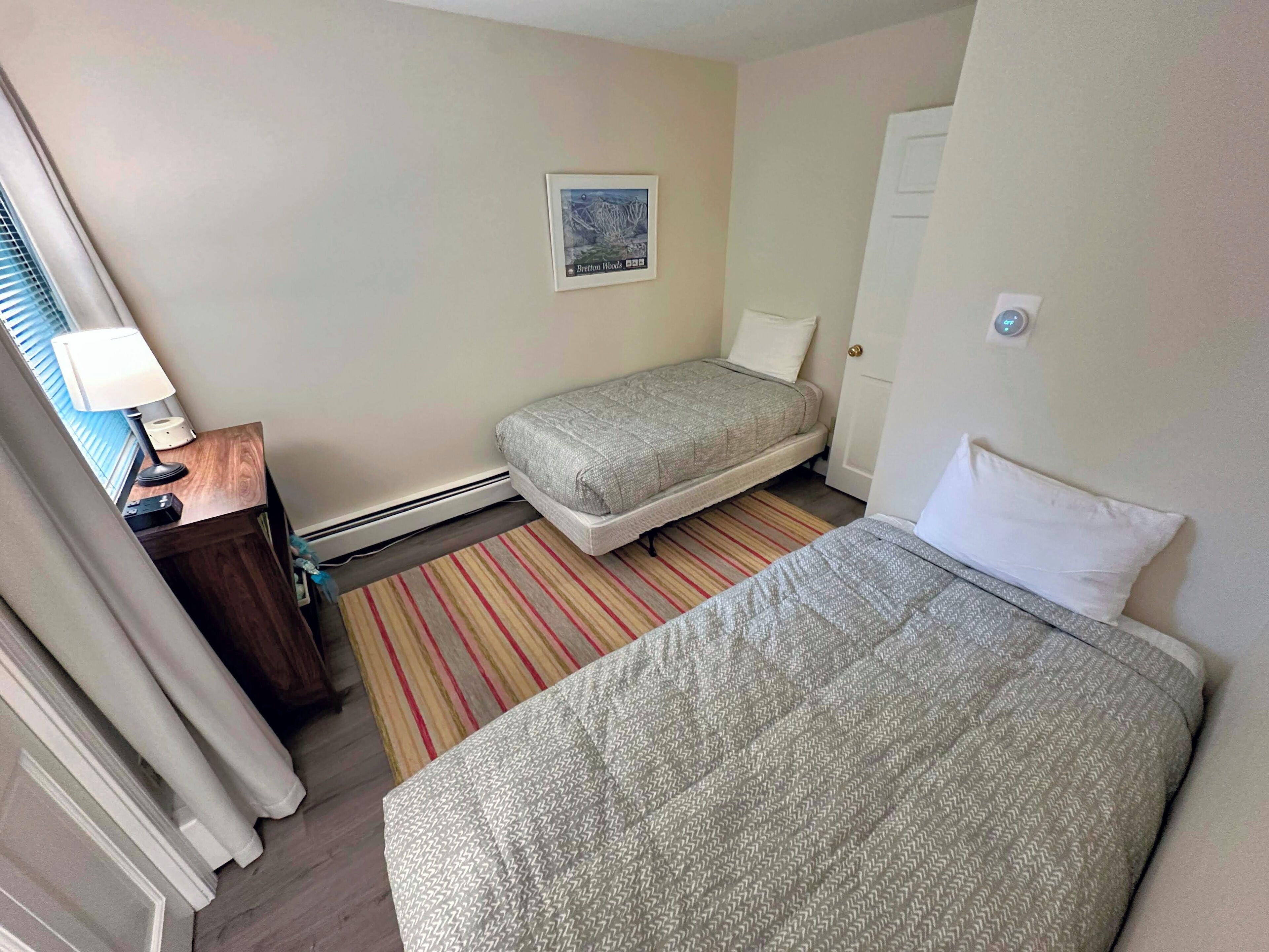 A hotel room with two beds, a desk with a lamp, a striped rug, and a framed picture on the wall.