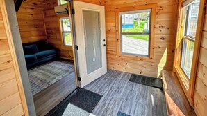 A wooden cabin interior with a door leading to a living area, a window with a view of a green landscape, and a doormat at the entrance. - Bmv7 Tiny Home Topline (Twin Mountain)