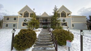 A multi-story residential building with a symmetrical facade, a central entrance with a staircase, and a snow-covered ground. - Sc10 Cozy Bretton Woods Ground Level Condo (Bretton Woods)