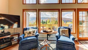A living room with two blue armchairs, a TV, and a chessboard on a small table. Large windows offer a view of mountains and trees. - SH4 Bretton Woods Condo With Hot Tub, Next to Skiing and Mt Washington Hotel, ski Slopes Views! (Bretton Woods)