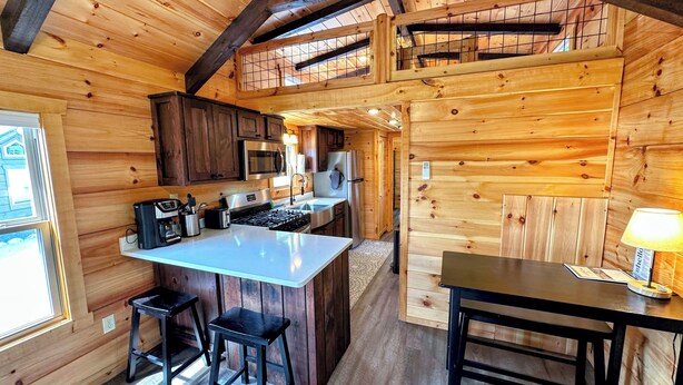 A wooden cabin with a kitchen featuring a central island, bar stools, and a dining table. - Bmv6 Tiny Home Topline (Twin Mountain)