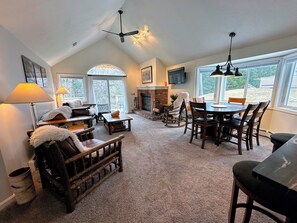 A cozy living room with a fireplace, a dining area, and a large window offering a view of the outdoors. - Fv44 Tastefully Updated Single Level With AC, Wifi, Minutes From Skiing! (Bretton Woods)