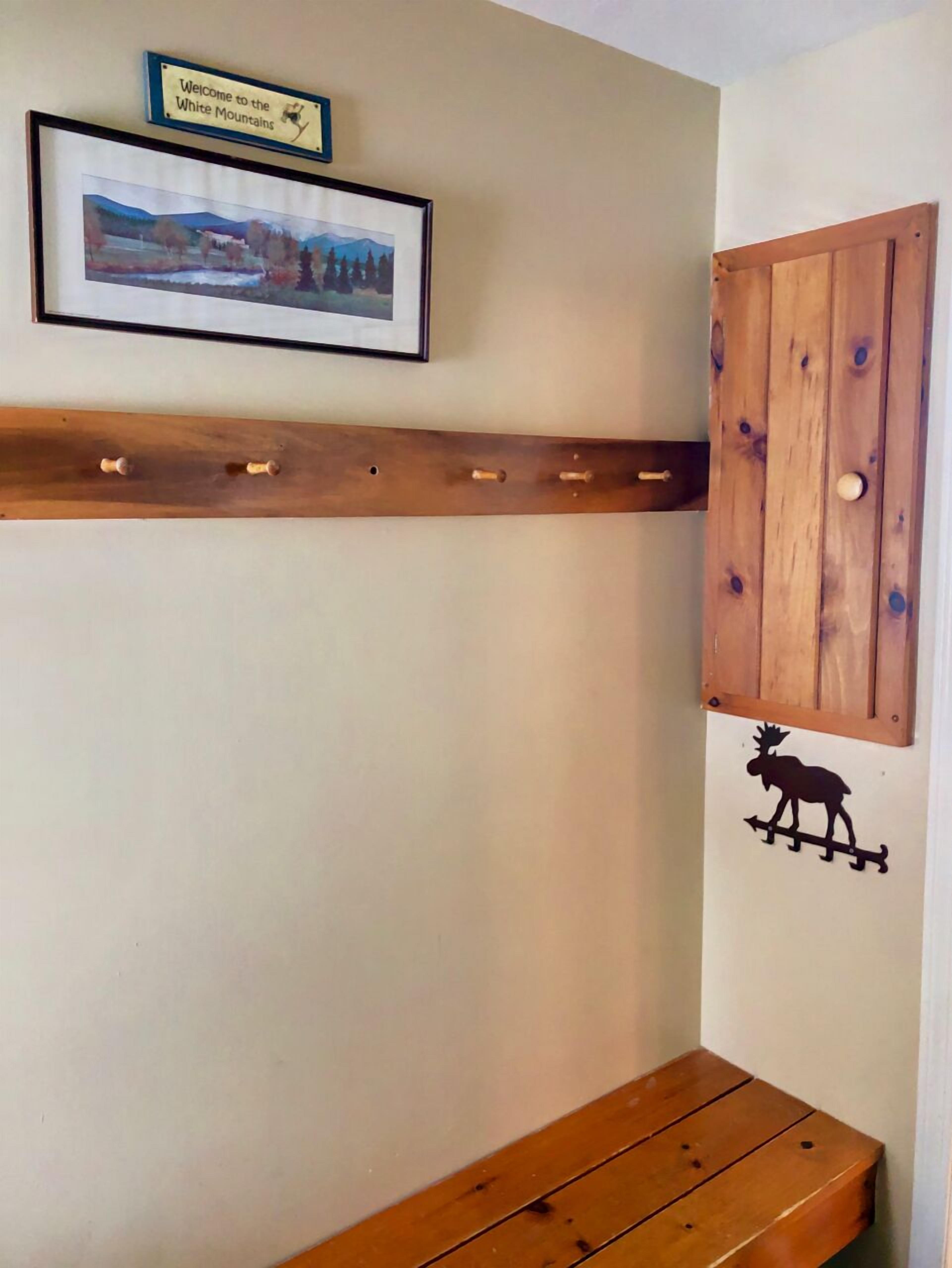 Una habitación con un banco de madera, un perchero montado, un cartel de bienvenida enmarcado y una decoración con silueta de alce.