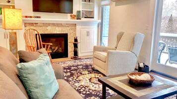 A living room with a fireplace, a television mounted above it, and a large window providing a view of the outdoors.