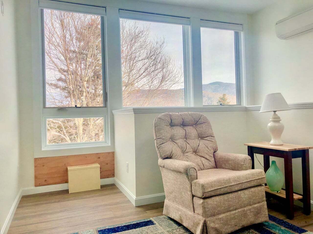 A room with a large window, a cushioned chair, a lamp, and a small table.