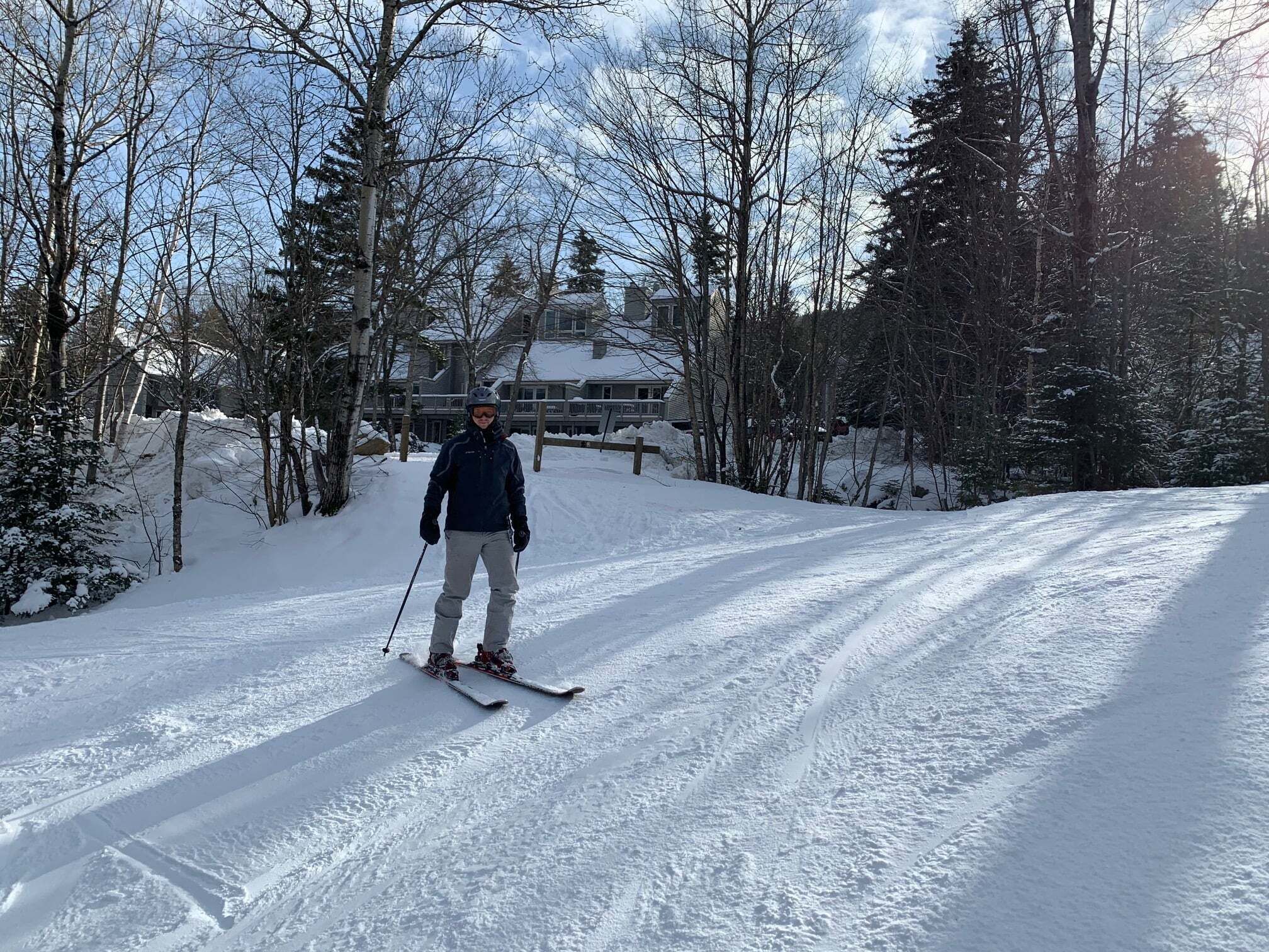 A person skiing on a snow-covered path with trees and a building in the background.