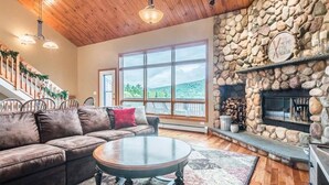 A cozy living room with a stone fireplace, a large window offering a scenic view, a wooden ceiling, and a comfortable seating area. - V3 Gorgeous Ski-in Mountain View Townhouse With Amazing Views in Bretton Woods (Bethlehem)