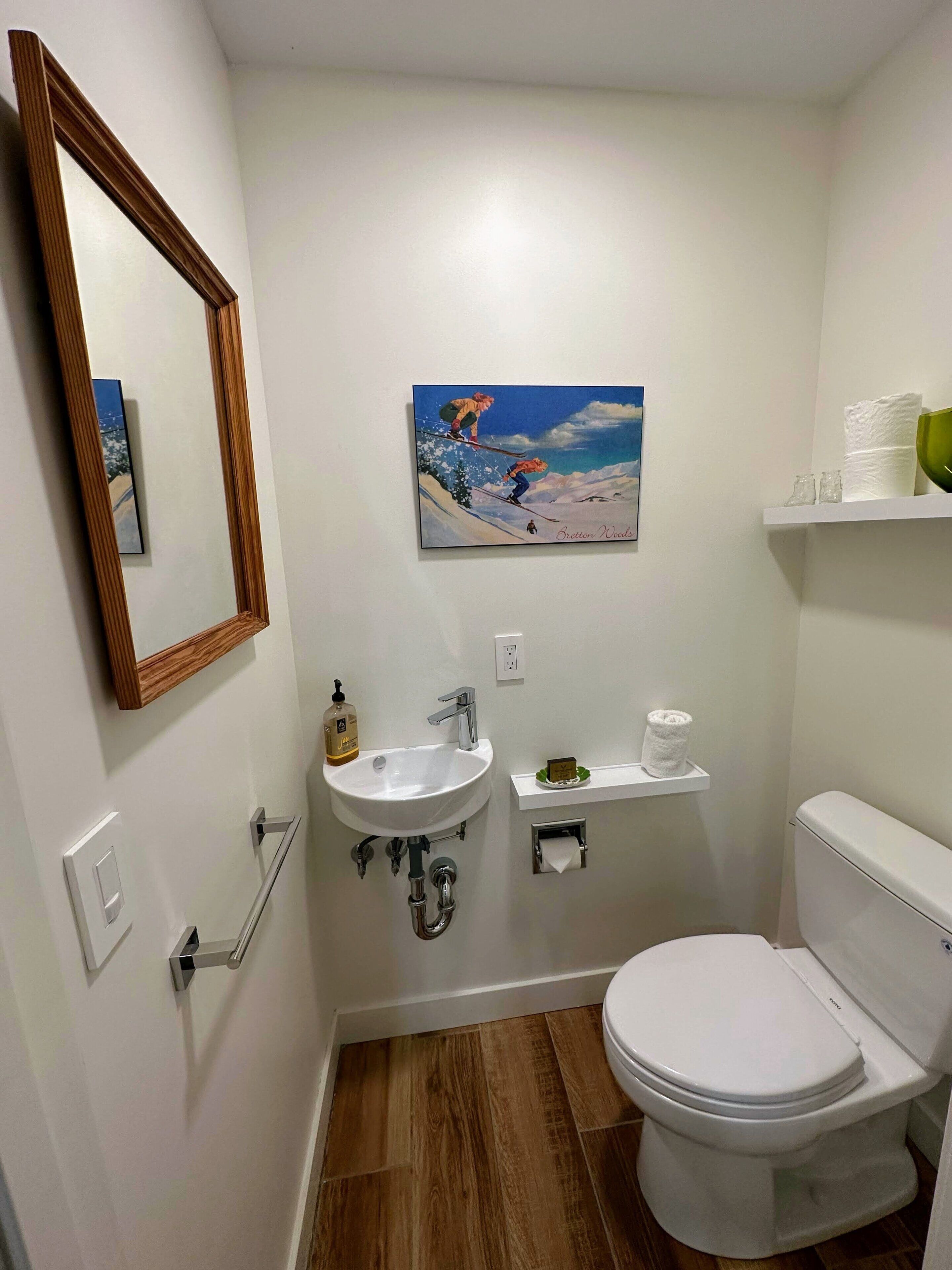Casa de banho com lavatório branco, sanita, espelho, quadro e prateleira com artigos de higiene pessoal.