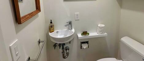 Casa de banho com lavatório branco, sanita, espelho, quadro e prateleira com artigos de higiene pessoal.