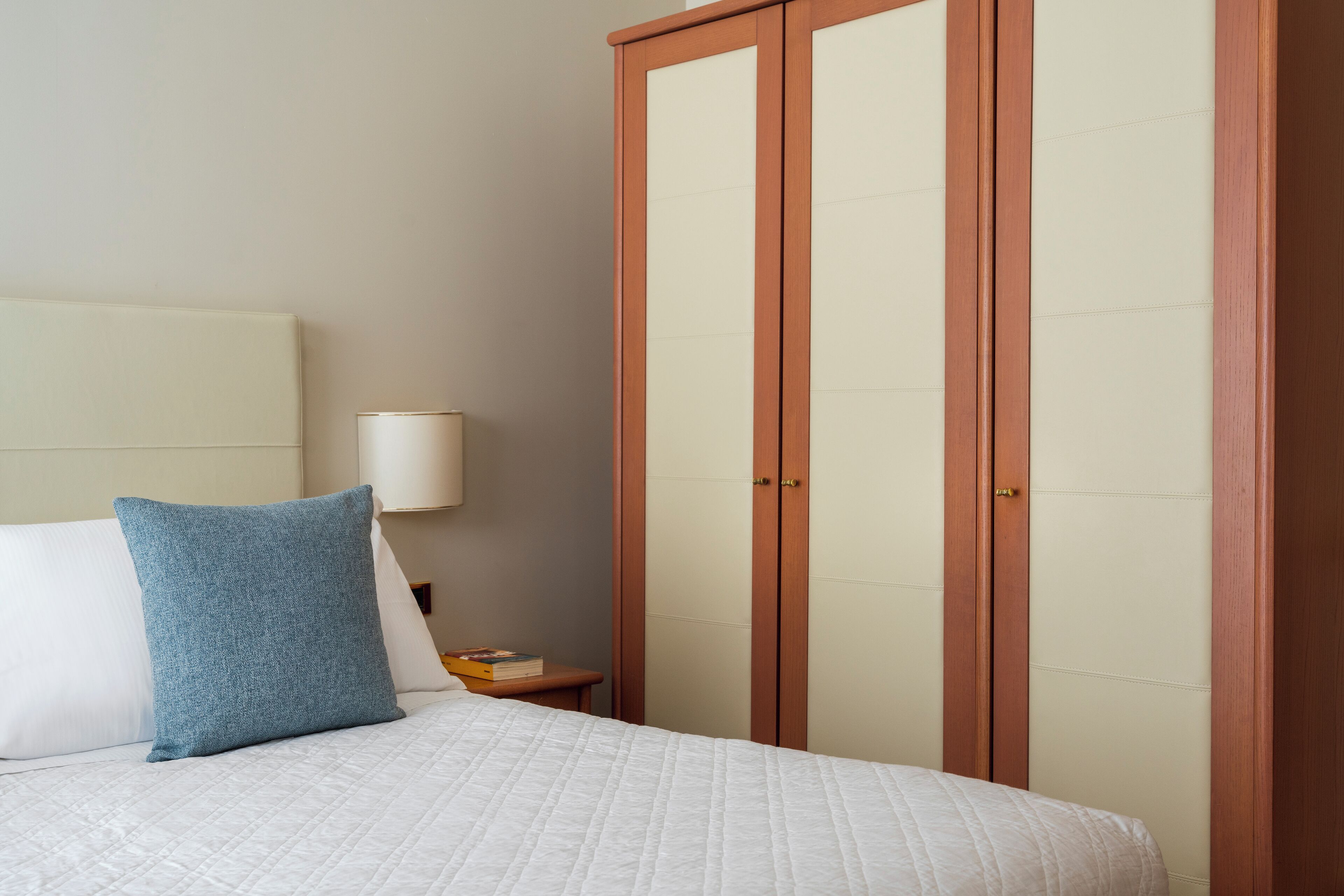 A neatly made bed with a blue pillow and a white quilted bedspread, a bedside table with a lamp, and a wooden wardrobe with two doors.