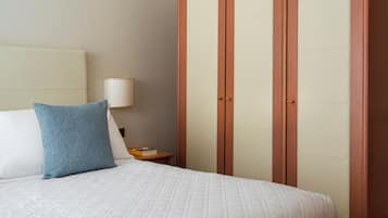 A neatly made bed with a blue pillow and a white quilted bedspread, a bedside table with a lamp, and a wooden wardrobe with two doors.