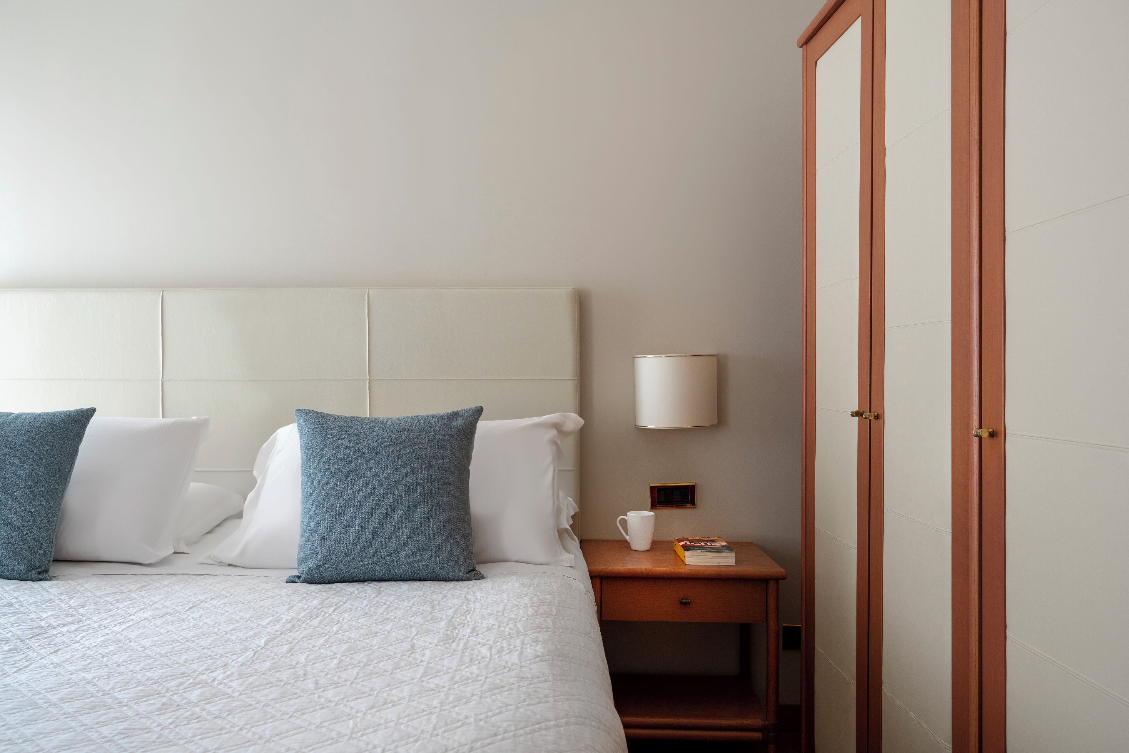 Una cama bien hecha con almohadas blancas y azules, una mesita de noche con una lámpara y un libro, y un armario de madera.