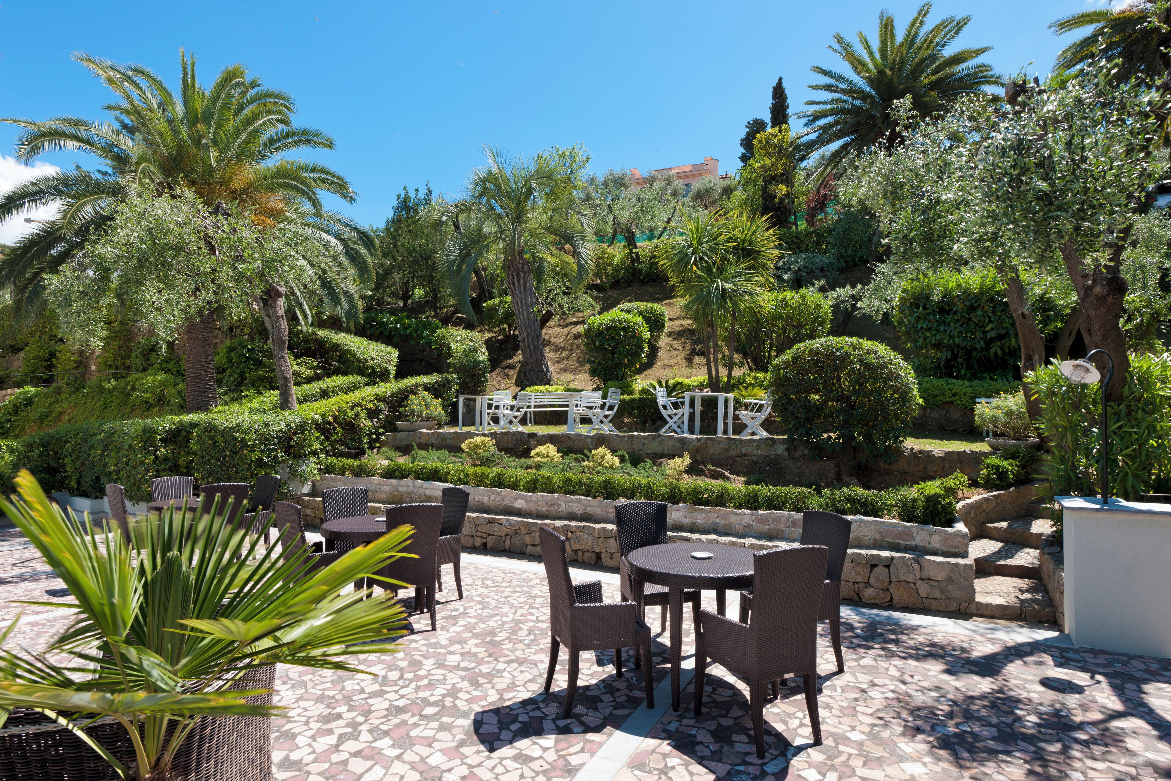 Un espace extérieur aménagé en terrasse, doté de mobilier en osier, d’un sol en pierre et d’une variété de plantes, notamment des palmiers et des arbustes.