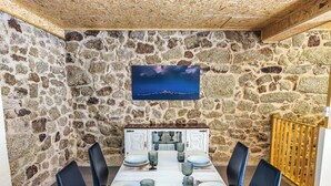 A dining area with a stone wall, a television mounted on the wall, and a table set for six with chairs. - FONTE DA ROSA GUEST HOUSE CENTRAL STUDIO (Belmonte)