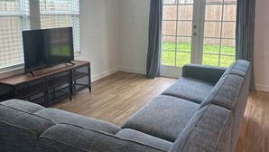 Ein Wohnzimmer mit einem großen, grauen Ecksofa, einem Flachbildfernseher auf einem Holzständer und einem Deckenventilator.
