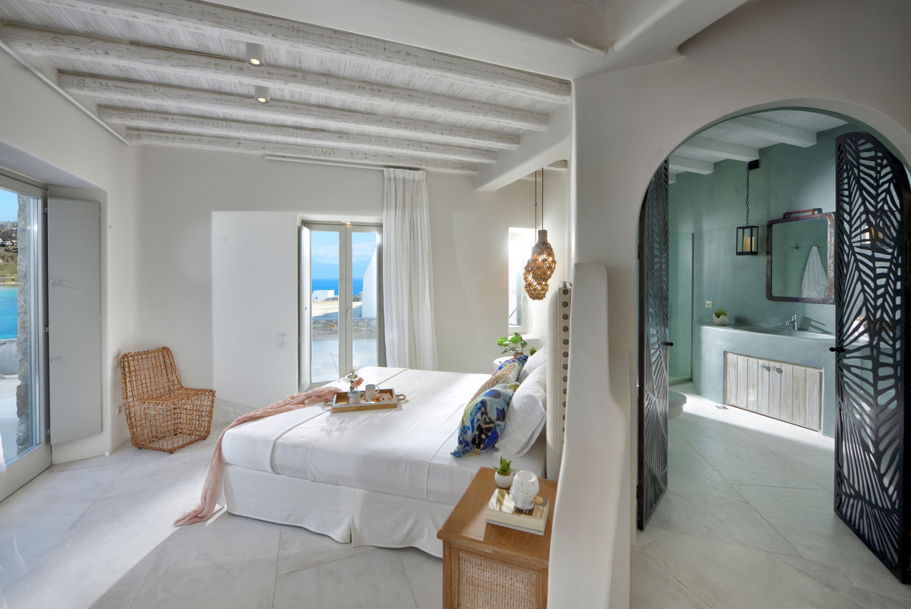 A spacious bedroom with a large bed, a wicker chair, a bedside table with a tray, and a view of the sea through a window.