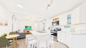 A modern kitchen with white cabinetry, a central island, a dining area with a table and chairs, and a living area with a sofa and TV.