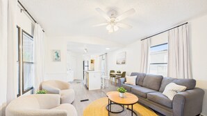 A modern living room with a grey sofa, a round coffee table, and a beige armchair. There is a kitchen area in the background with a dining table and chairs.