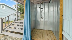 A covered outdoor shower area with a wooden deck and steps leading to a light blue house.