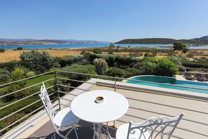 Ein Balkon mit weißem Tisch und Stühlen, der Blick auf einen Pool und eine malerische Landschaft mit Hügeln und einem Gewässer freigibt.