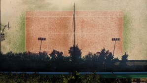 Ilmakuva tenniskentästä, jossa on verkko, ja jonka ympärillä on puita ja vihreä aita.