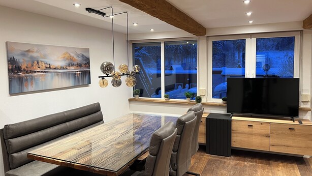 Een moderne eethoek met een houten tafel en lederen stoelen, een schilderij aan de muur en een sneeuwlandschap te zien door grote ramen.