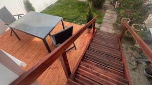 Een houten terras met een tafel en stoelen, omringd door een witte schutting en begroeiing.