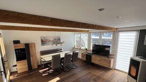 Een moderne woonkamer met een eethoek, houten vloeren, een open haard en een groot raam met zonwering.