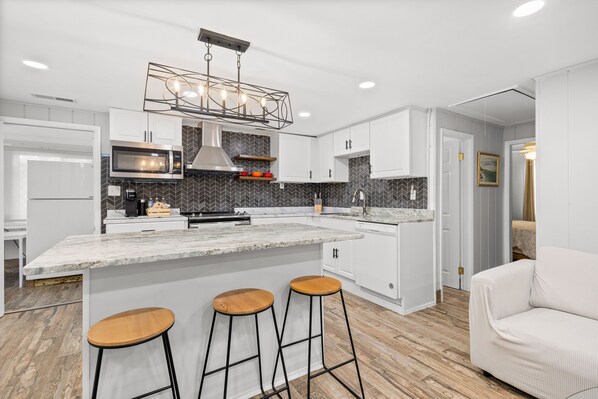 A modern kitchen with white cabinets, a large island with stools, a built-in microwave, and a unique pendant light.