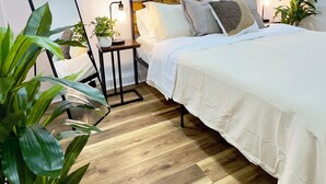 A modern bedroom with a wooden floor, a bed with white bedding, a bedside table with a lamp, a mirror, and a wall art piece. - Cozy 1BR in Philly with Rain Shower (Philadelphia)