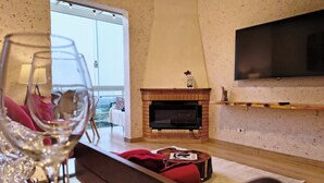 Ein Wohnzimmer mit Kamin, einem an der Wand befestigten Fernseher und Blick ins Freie durch eine Glastür.