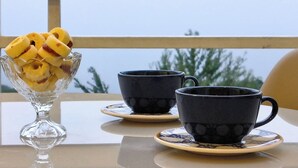 Ein Tisch mit einer Glaskugel voller Obst, zwei schwarze Tassen mit Untertassen und Blick auf Bäume durch ein Fenster.
