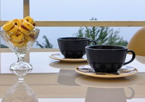 A table with a glass bowl of fruit, two black cups with saucers, and a view of trees through a window. - Duplex apartment with beautiful views. (Campos do Jordão)