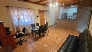 Un salon avec un canapé en cuir noir, une table à manger avec des chaises, un coin cuisine avec un mur en briques et une fenêtre ornée de rideaux légers.