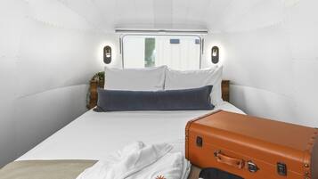 A compact hotel room with a bed, pillows, a suitcase, and a wall-mounted light fixture.