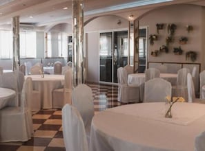 Uma sala de banquetes com mesas e cadeiras redondas, janelas amplas e um chão quadriculado.