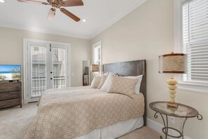 A bedroom with a bed, a ceiling fan, a table with a lamp, and a view of the beach through the door. - Villa Palmeira (Santa Rosa Beach)